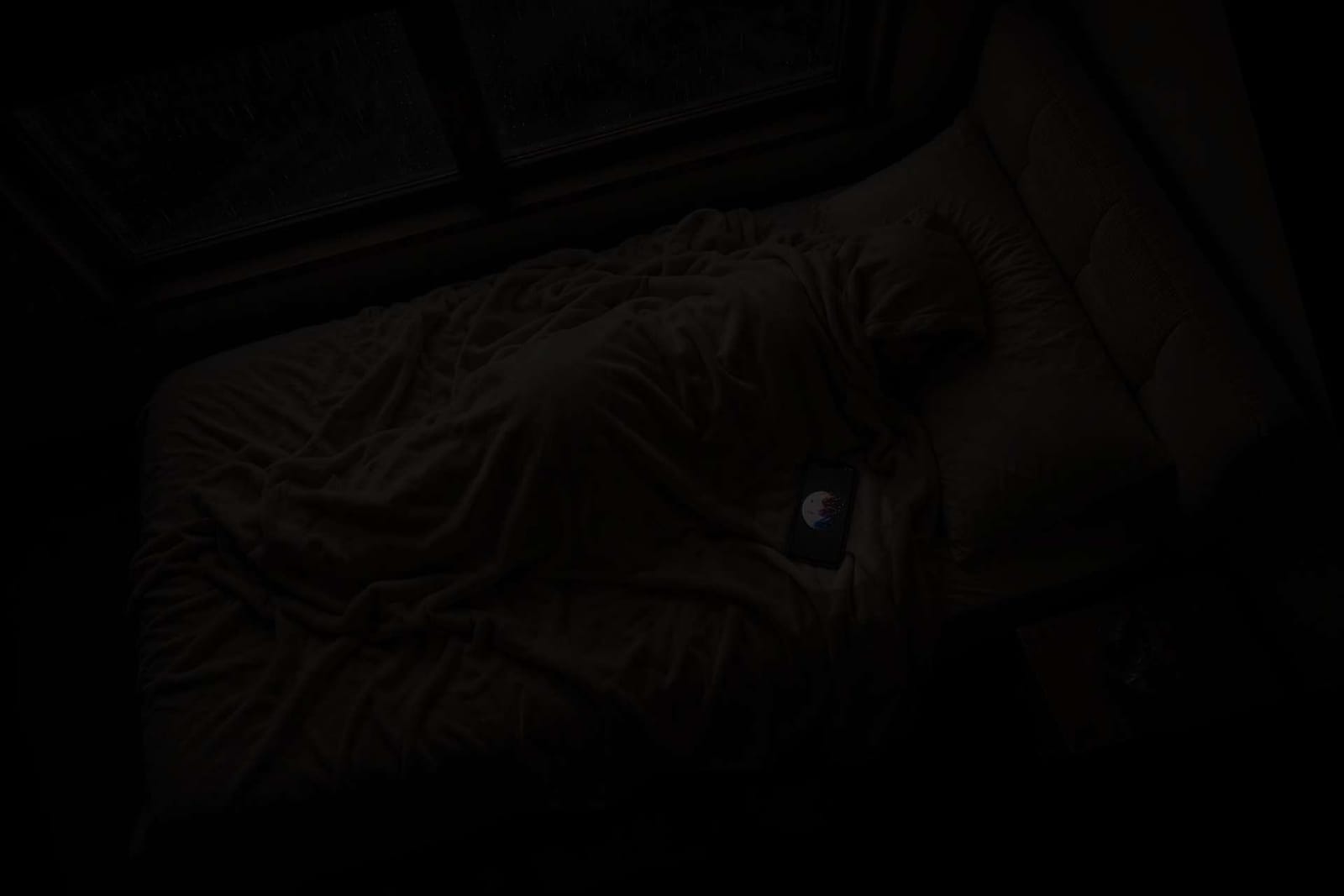 A dark bedroom at night with a phone glowing alone on an unmade bed