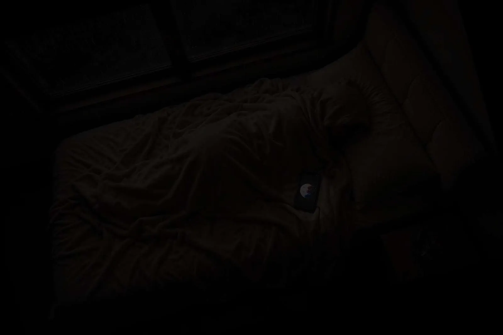 A dark bedroom at night with a phone glowing alone on an unmade bed